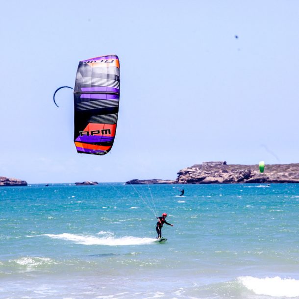 Female Kitesurfer riding toeside towards the shore with a pink, violet and grey kite on turquoise water and other kites on the back during the Kite Sisters Women Kiteboarding Camp in Essaouira Morocco
