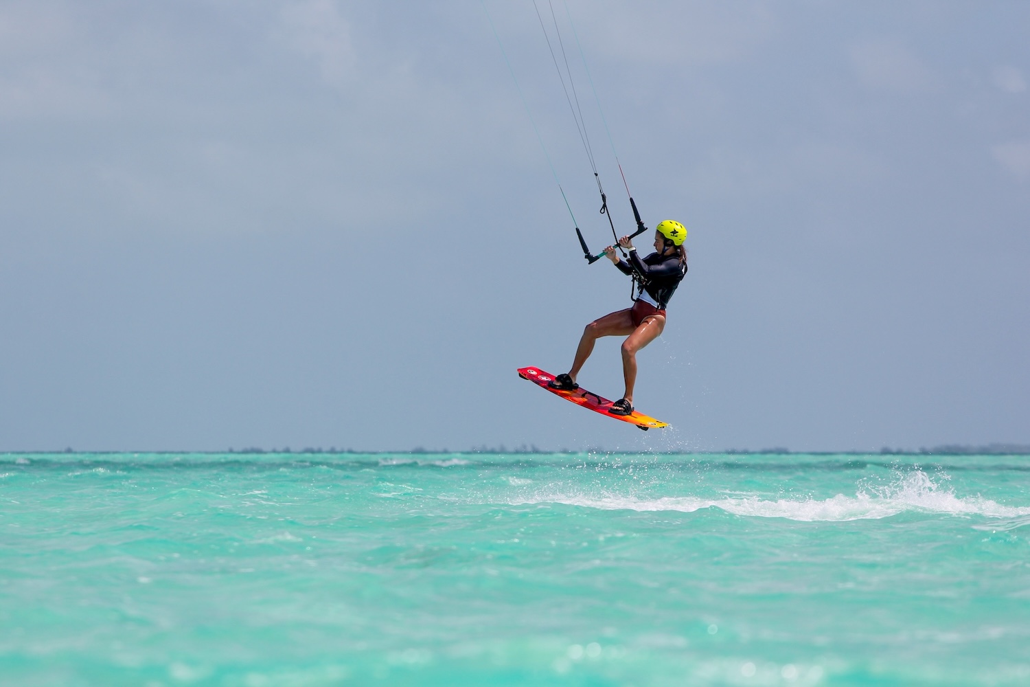 Kiteboarder in a black wetsuit with a yellow helmet leaps above turquoise water, gripping the kite bar and lines.
