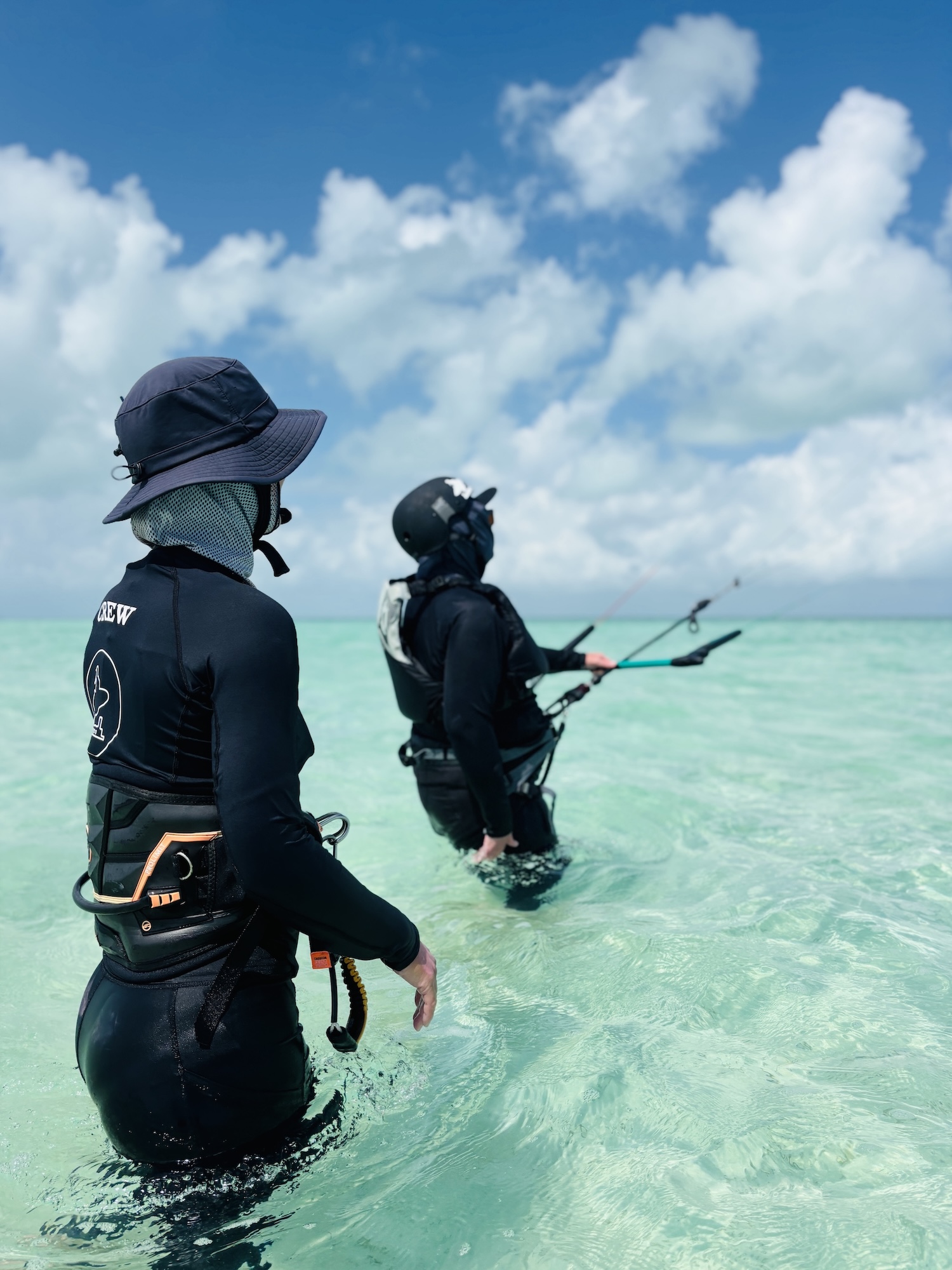 Two water sports enthusiasts stand waist-deep in turquoise water, wearing black wetsuits, harnesses, and protective headgear under a bright sky.