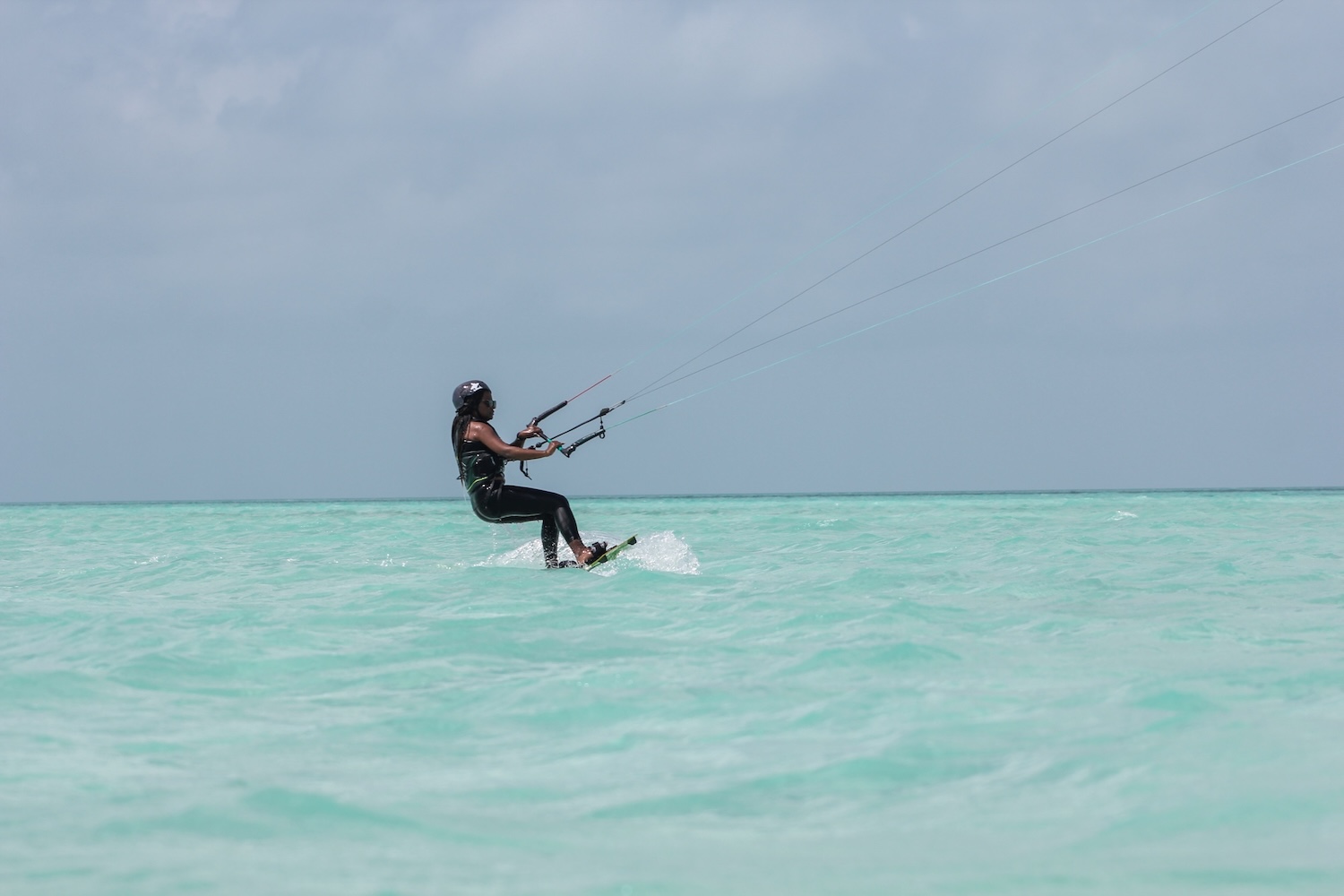 Kiteboarder riding turquoise ocean water, wearing a helmet and wetsuit, gliding with a kite overhead.