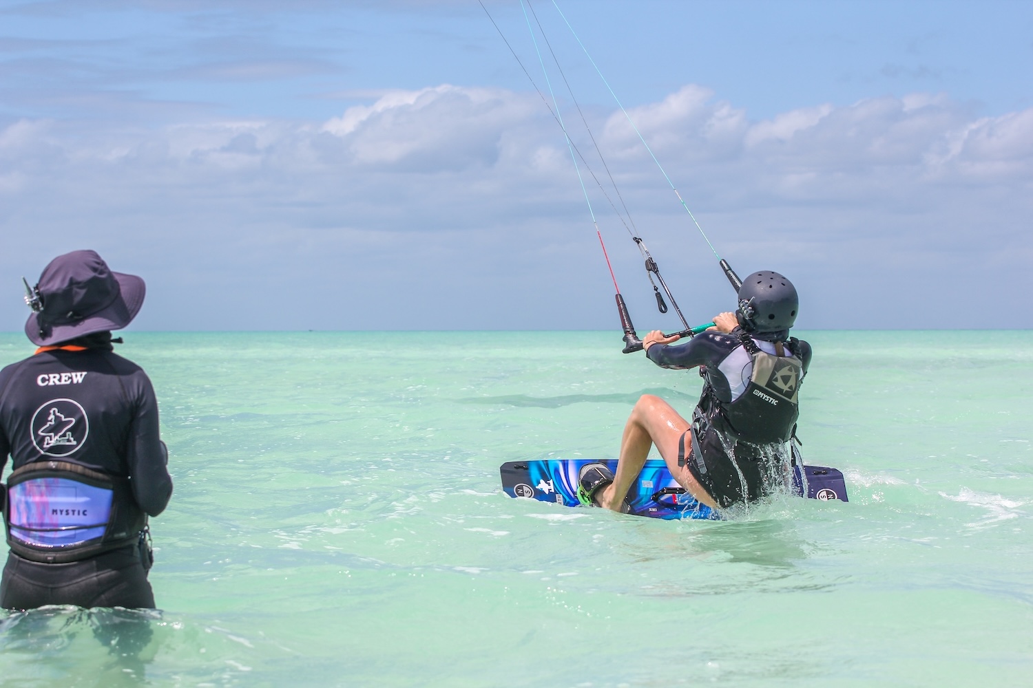 Kiteboarder in a helmet and harness skims the turquoise water while a crew member watches from shallow water nearby at the beach.