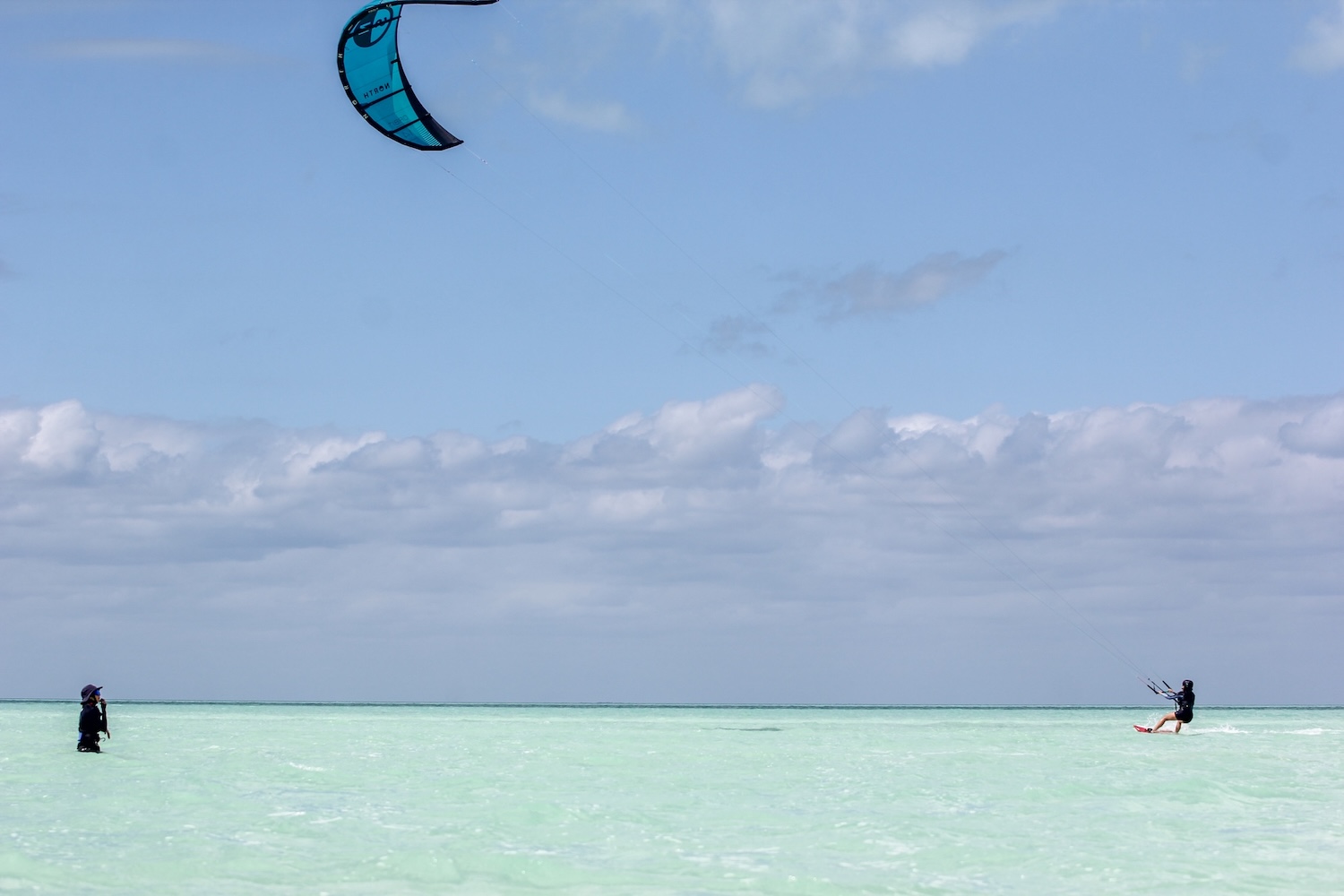 Two people in shallow turquoise water: a kitesurfer slides across the right while another person stands waist-deep on the left; blue sky and clouds above
