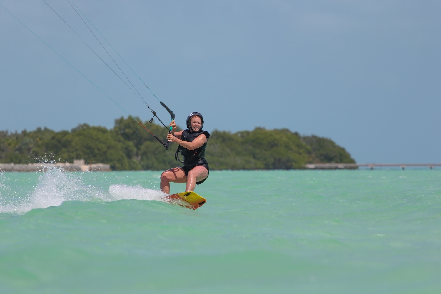 Kiteboarder riding a yellow board across turquoise water, wearing a helmet and life vest, with lines overhead and a green shoreline in the distance.