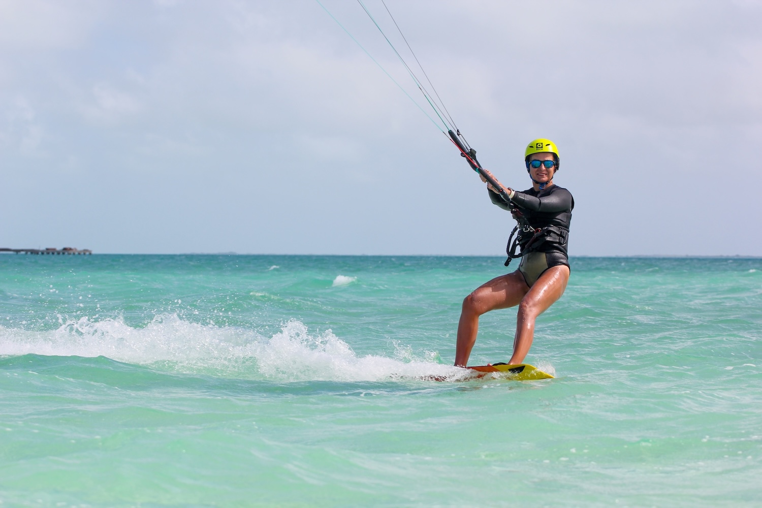 Kitesurfer in a black wetsuit and yellow helmet rides a board on turquoise water, holding the control bar.