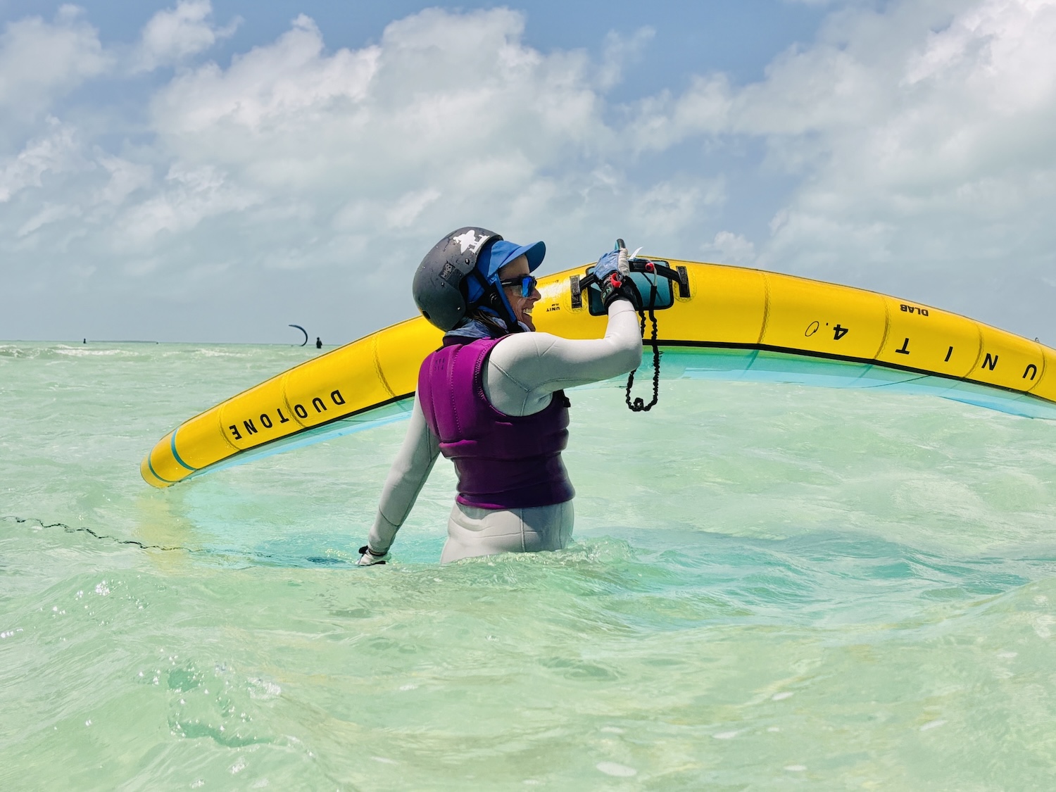 Person wearing a helmet and purple life vest stands in shallow turquoise water, lifting a bright yellow windsurf board over their shoulder.