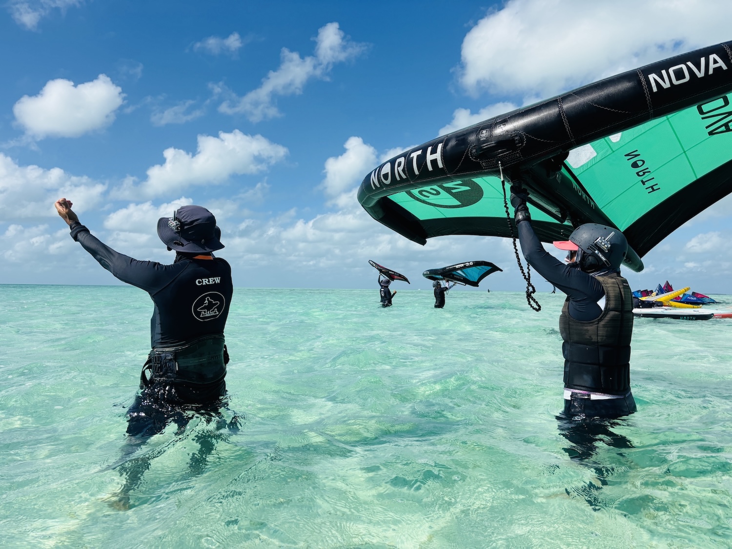 Windsurfing crew in turquoise water raise bright sails, preparing to launch under a blue sky.