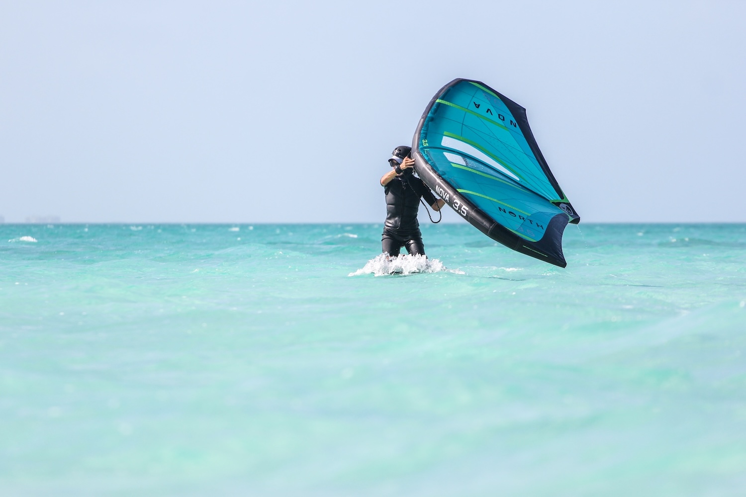Kitesurfer in a black wetsuit standing in shallow turquoise water, holding a large blue-green kite