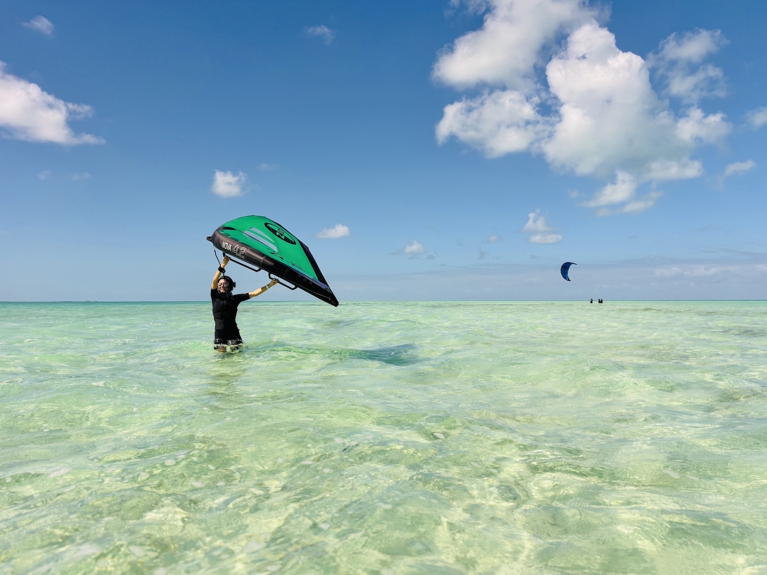Person standing in clear shallow water holding a green and black kite above their head, ready to kiteboard on a sunny day.