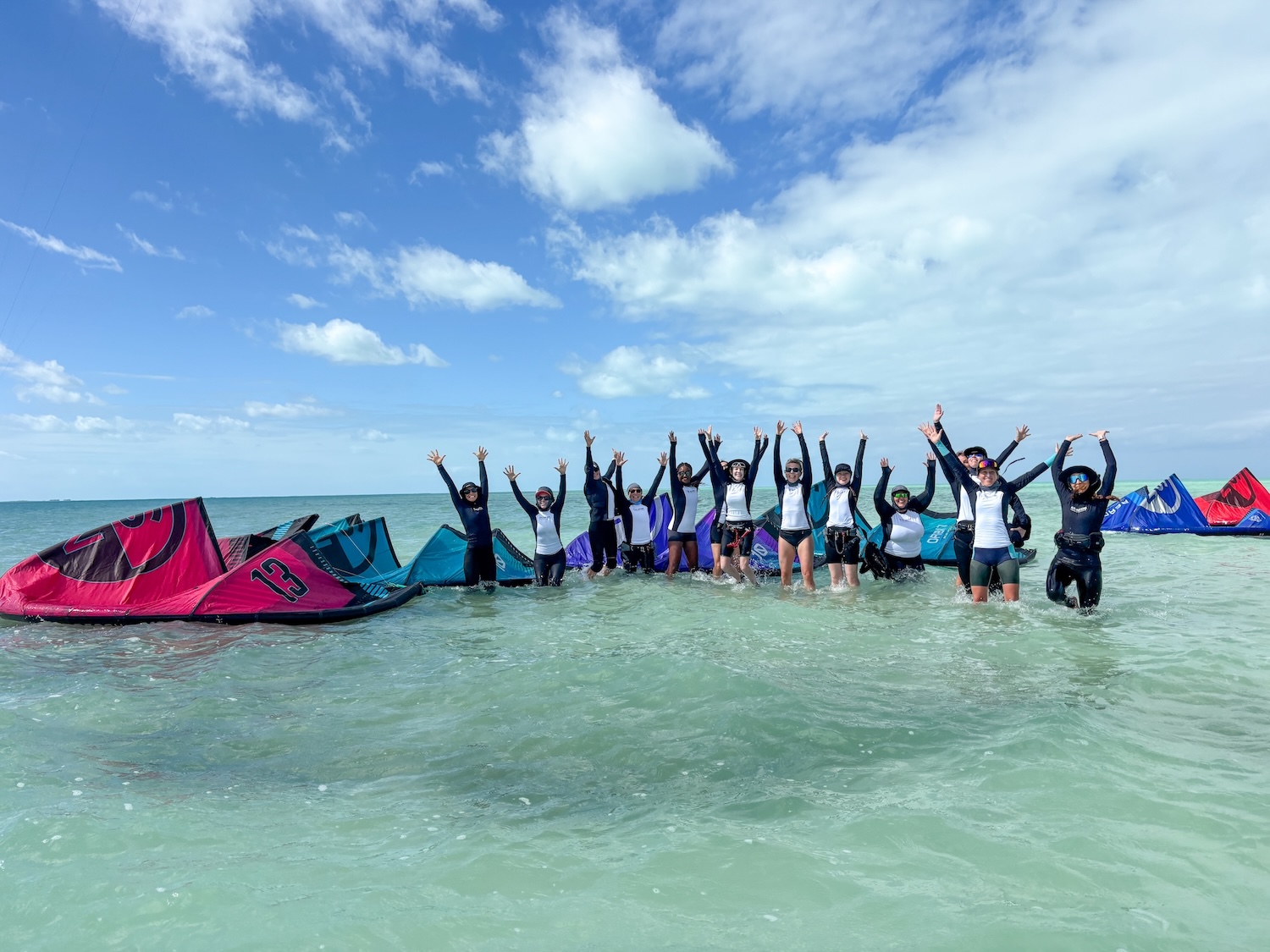 Group of people in wetsuits standing waist-deep in turquoise water with kites on the shore, arms raised in celebration.
