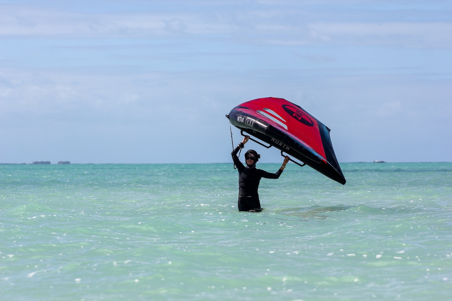 Kitesurfer in a wetsuit stands waist-deep in turquoise water, lifting a red and black kiteboard above their head as waves crash nearby