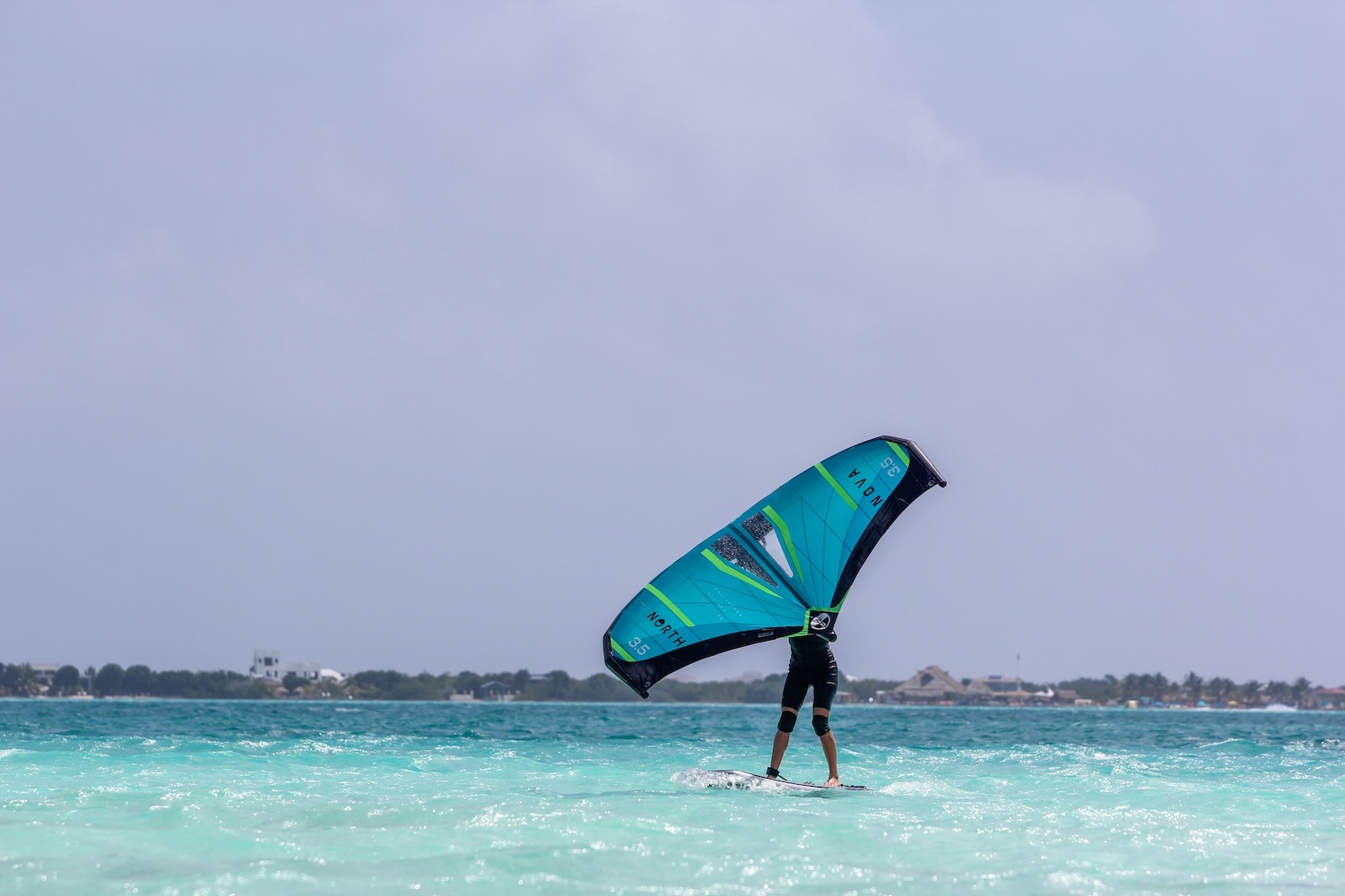 Windsurfer gliding on turquoise shallow water with a bright teal sail near a distant shoreline.