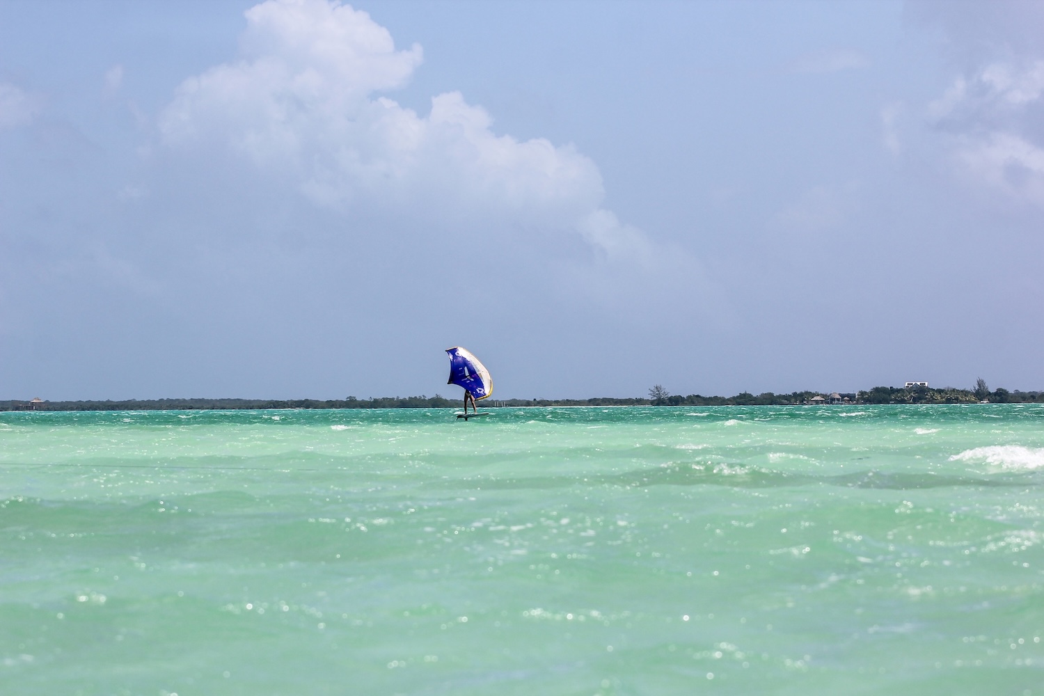 Lone windsurfer on turquoise water with a blue sail, near a distant shoreline under a cloudy sky.