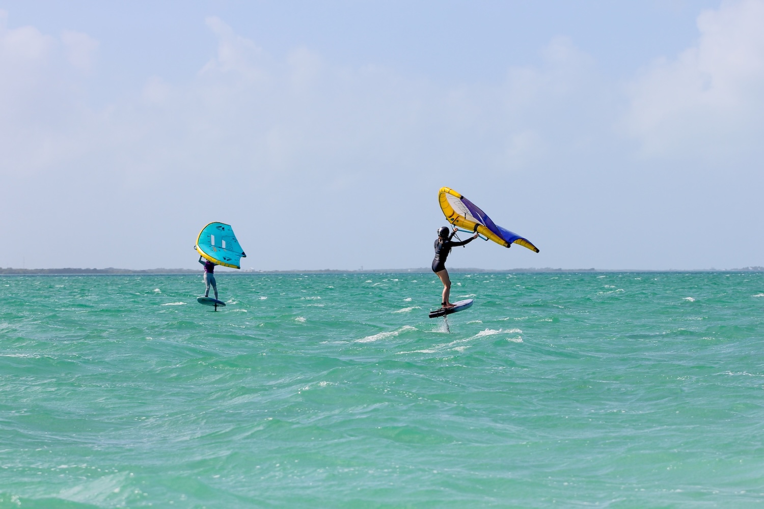 Two kiteboarders ride the turquoise ocean, one with a teal wing and the other with a yellow‑and‑purple wing, under a blue sky.