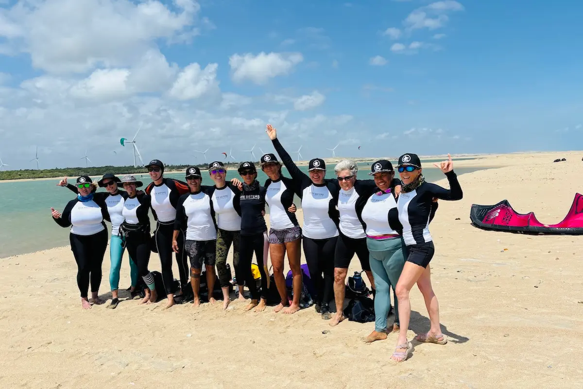 Group of women in Kite Sisters rash guards posing happily on a sandy beach with kites and wind turbines during a kiteboarding retreat in Brazil.