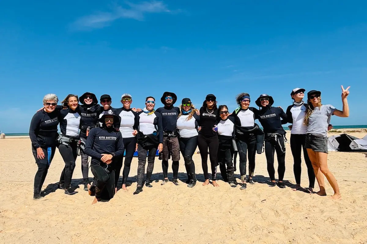 Large group of women and instructors in Kite Sisters gear posing together on a sandy beach under a blue sky during a Morocco kitesurf retreat in Dakhla.