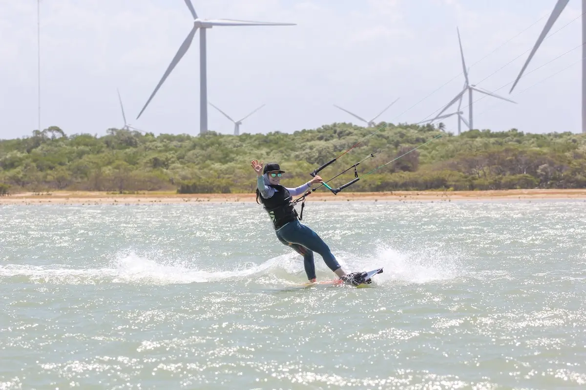 Kite Sisters woman waving and smiling while kiteboarding with wind turbines in the background at Brazil retreat.