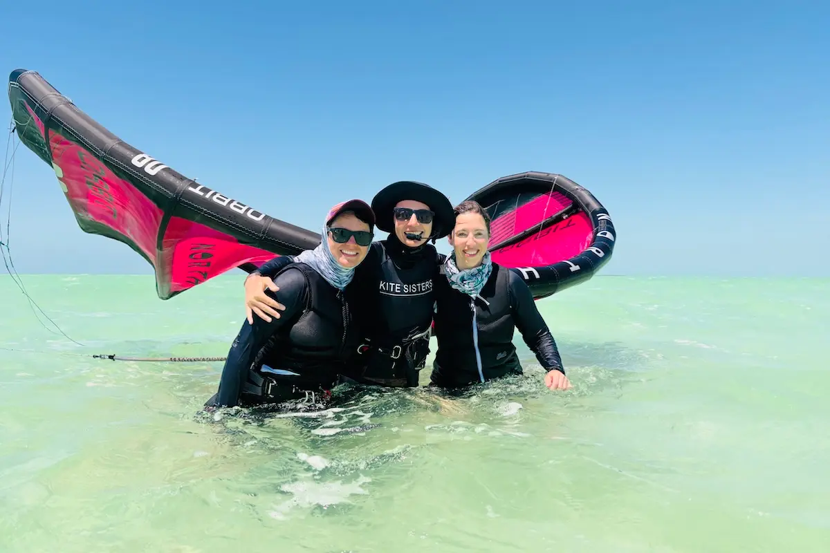 Kite Sisters instructor and two students smiling together with a kite in turquoise water in Belize.