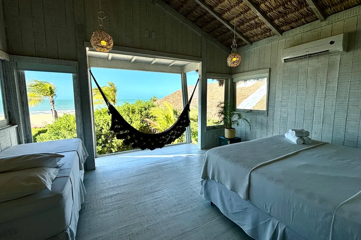 Bright beachside bedroom with a black hammock hanging between walls, white bedding, and a view of palm trees and the ocean through large windows