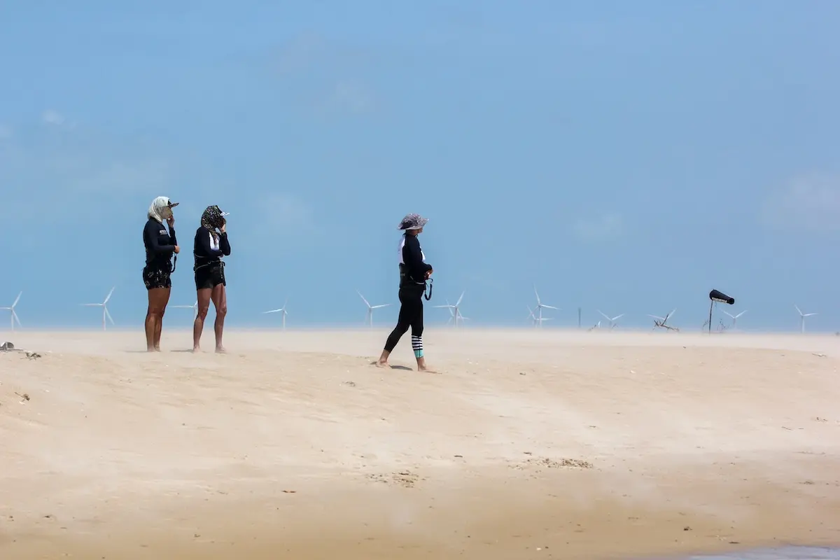 Three Kite Sisters women walking on a windy beach with wind turbines in the background at Brazil kitesurf retreat.