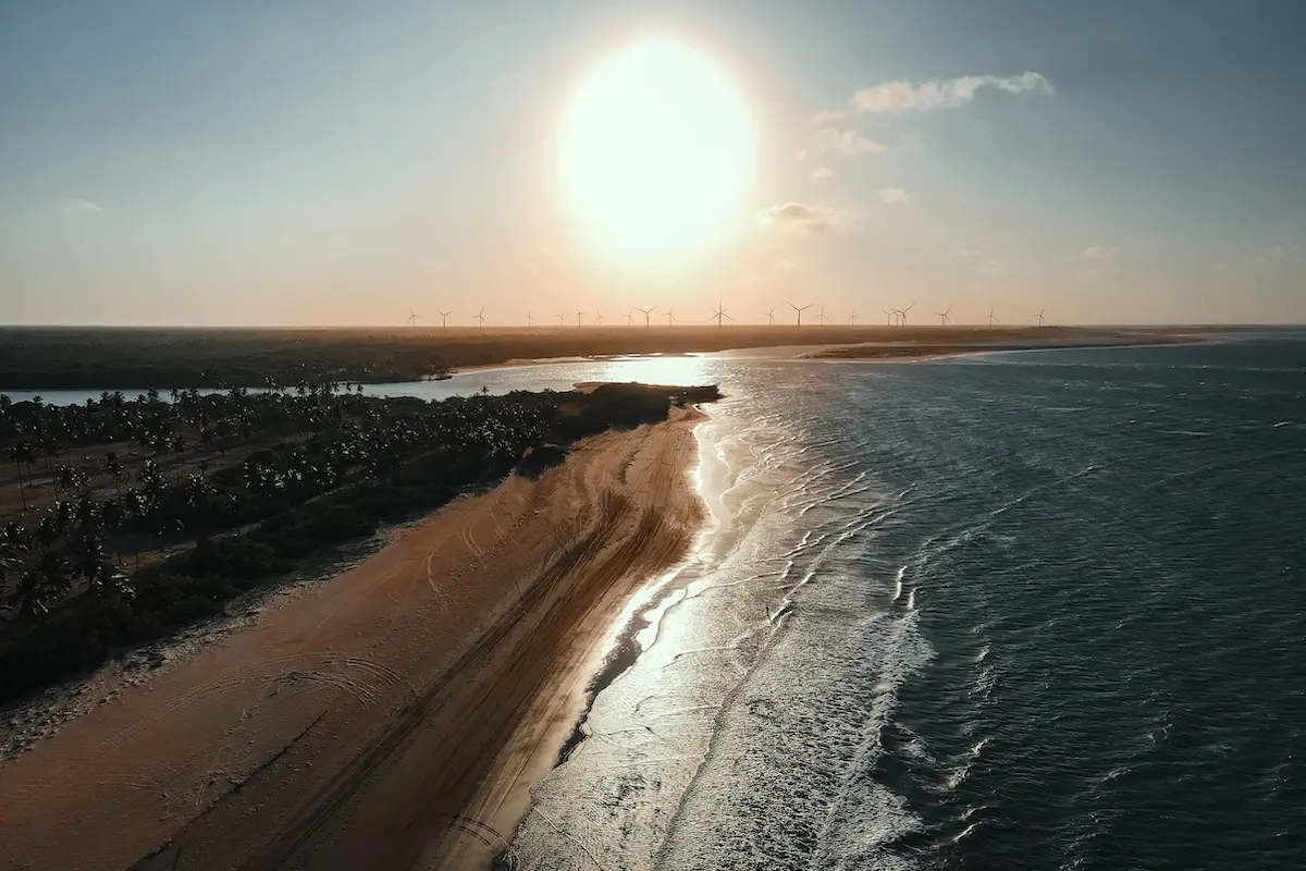 Aerial drone shot of a Brazilian coastline at golden sunset with wind turbines and palm trees, setting for a kitesurf retreat in Brazil.