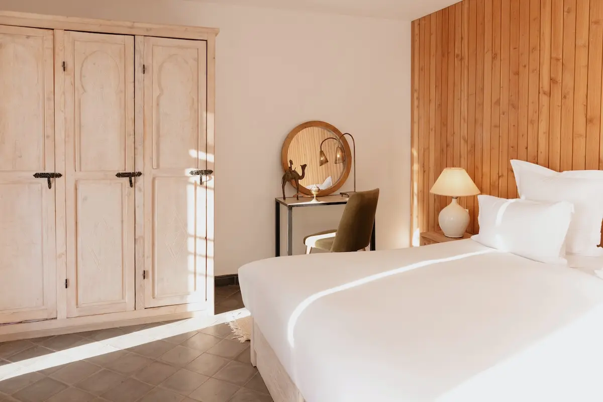 Bright and elegant bedroom with Moroccan wooden wardrobe, round mirror and white bedding at a Morocco kiteboarding retreat accommodation.