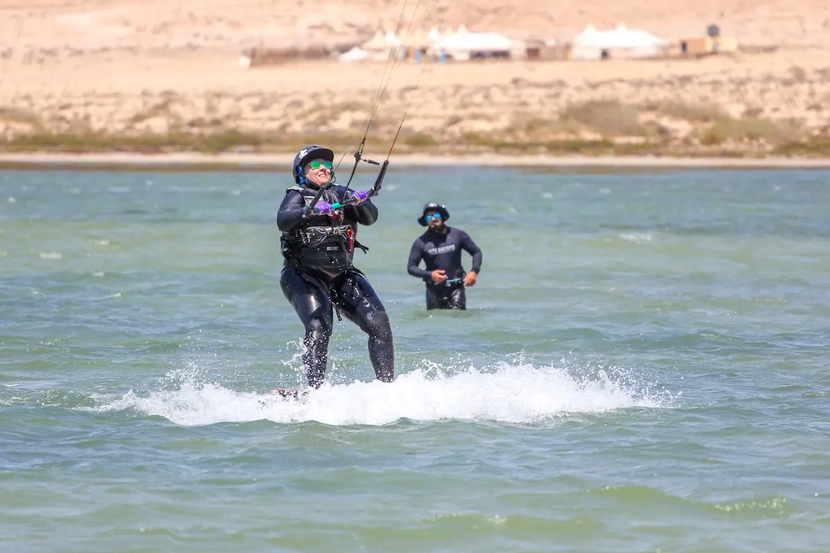 Smiling woman kiteboarding on the Dakhla lagoon with a Kite Sisters instructor watching in the background during a Morocco kiteboarding retreat.