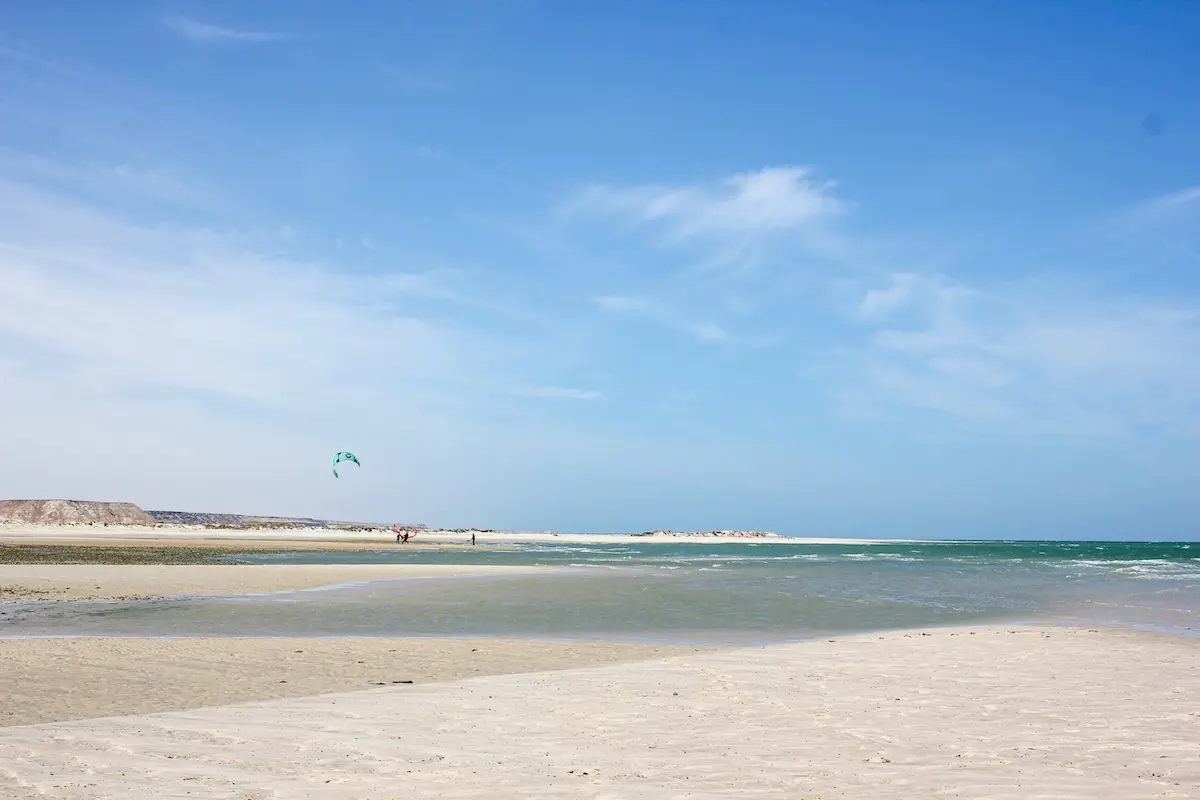 Wide white sand beach with turquoise water and a lone green kite flying in the blue sky at a Morocco kiteboarding retreat in Dakhla.