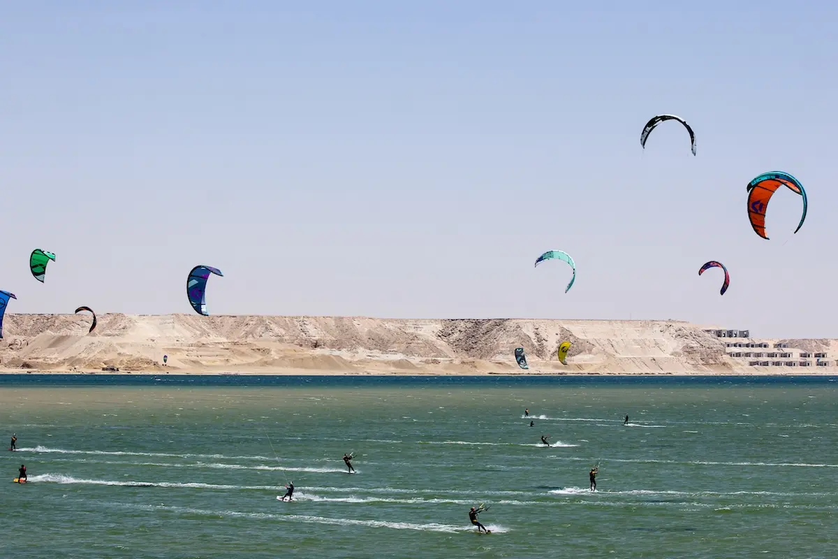 Wide shot of multiple kitboarders riding across the Dakhla lagoon with colorful kites filling the sky and desert cliffs in the background during a Morocco kiteboarding retreat.