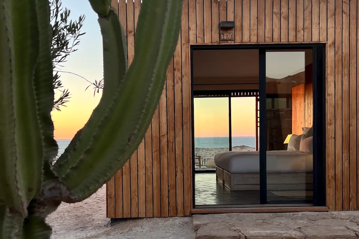 Wooden beachfront lodge with cactus in foreground and a warm ocean sunset reflected in the glass door at a Morocco kiteboarding retreat.