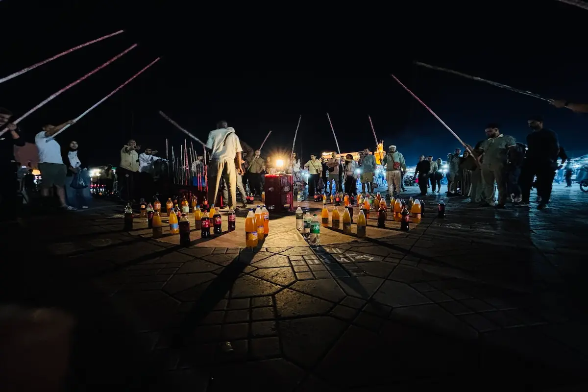 Night street performance with glowing bottles arranged on the ground at Djemaa el-Fna square, Marrakech, during a Morocco kiteboarding retreat cultural excursion.