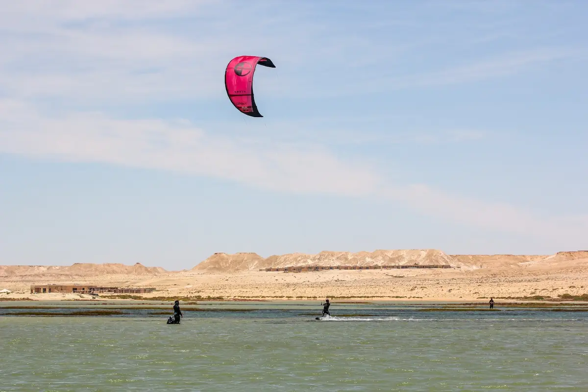 Large pink kite soaring above the Dakhla lagoon with three riders on the water and the retreat lodge visible in the desert background during a Morocco kiteboarding retreat.