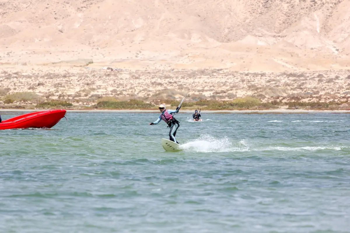 Woman riding a white surfboard strapless on the Dakhla lagoon with a second rider and desert hills in the background during a Morocco kiteboarding retreat.