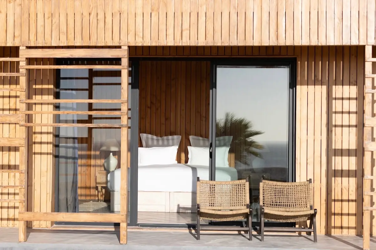 Wooden terrace with rattan chairs outside a bright bedroom with white bedding at a Morocco kitesurf retreat.