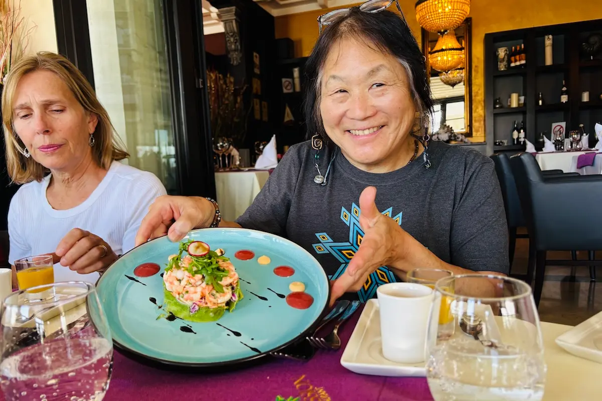 Two women at a restaurant table, one smiling with a thumbs up over a beautifully plated seafood dish during a Morocco kitesurf retreat.