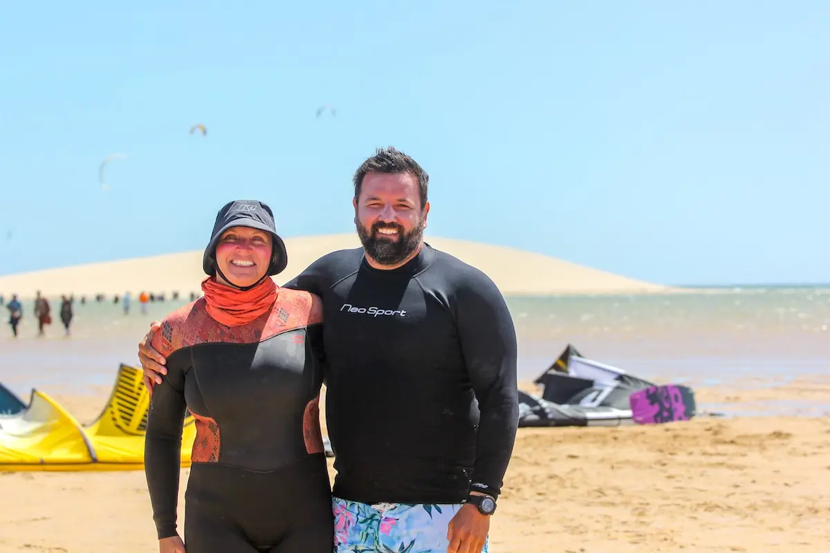 Smiling couple posing together on Dakhla beach with kites and a sand dune in the background during a Morocco kitesurf retreat.