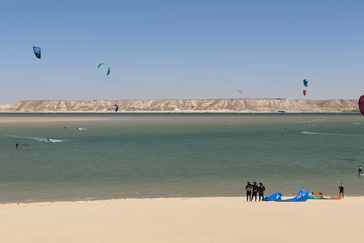 Wide view of the Dakhla lagoon with multiple colorful kites in the sky, riders on the water and white cliffs in the background during a Morocco kitesurf retreat.