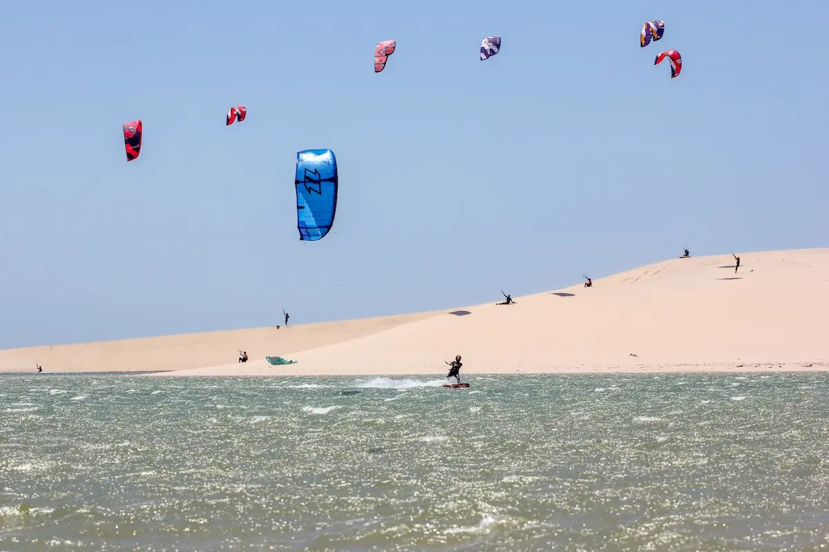 Multiple colorful kites flying above a large sand dune with kitesurfers riding the lagoon below during a Morocco kitesurf retreat in Dakhla.