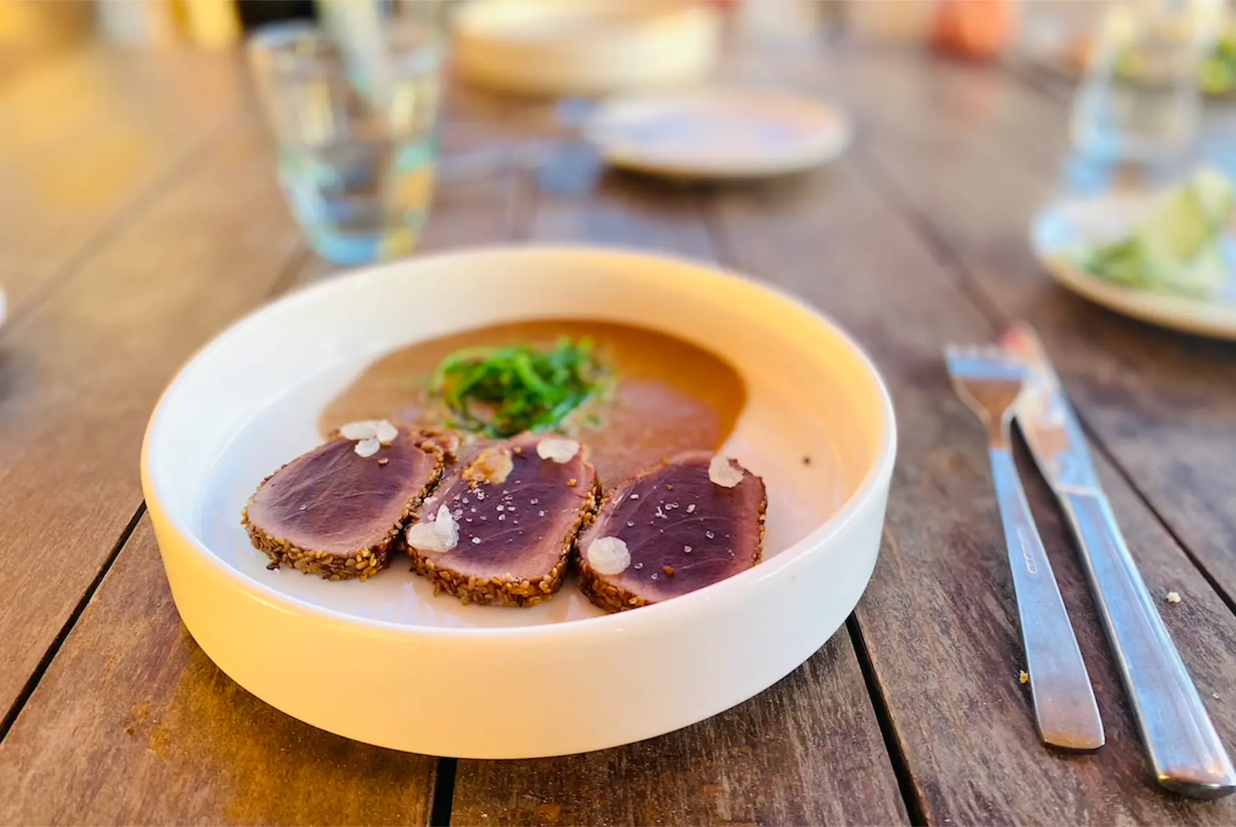Sliced sesame-crusted tuna with greens in a white bowl on a wooden table