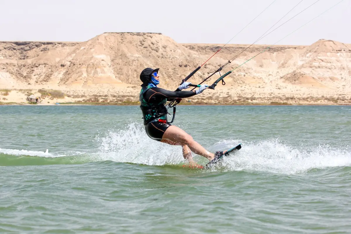 Woman in a black wetsuit carving hard on her kiteboard across the Dakhla lagoon with desert hills behind her during a Morocco kitesurf retreat.