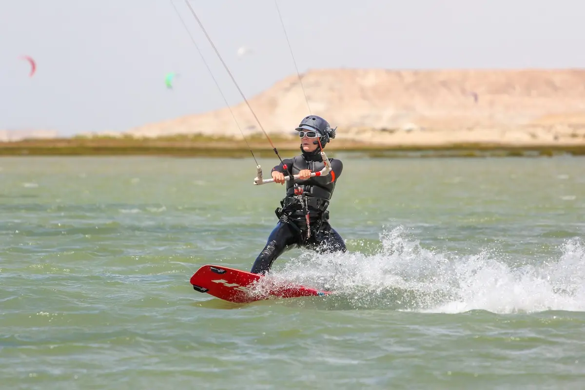 Woman in a black wetsuit riding a red kiteboard across the Dakhla lagoon with desert hills in the background during a Morocco kitesurf retreat.