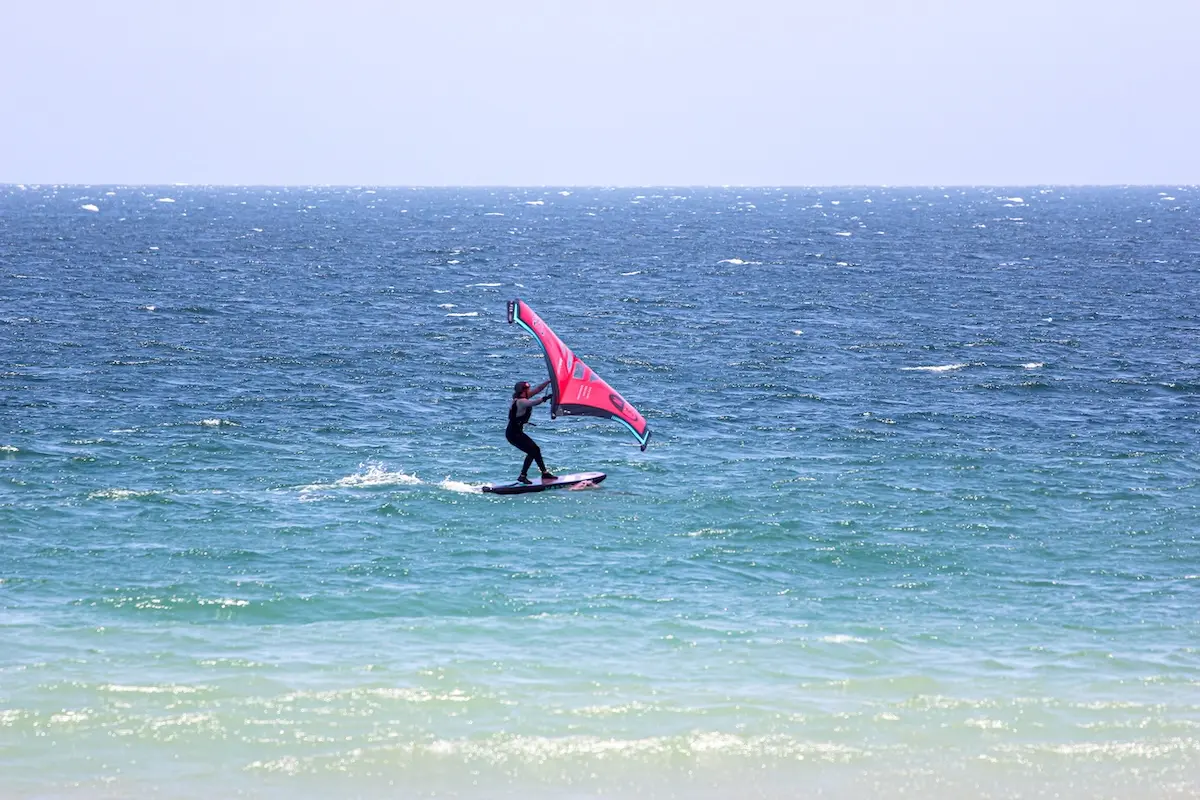 Woman wingfoiling on a foil board holding a red wing over turquoise ocean waters at a women's wingfoil camp in Peru.