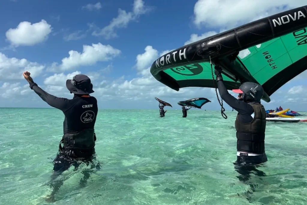 Kite Sisters crew member raising a fist while a group of women practice wingfoiling with North Nova wings in turquoise Belize water during a women's wingfoil retreat.