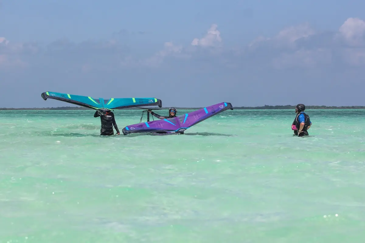 Three women practicing wing handling with colorful teal and purple wings in clear shallow Belize water during a women's wingfoil retreat.