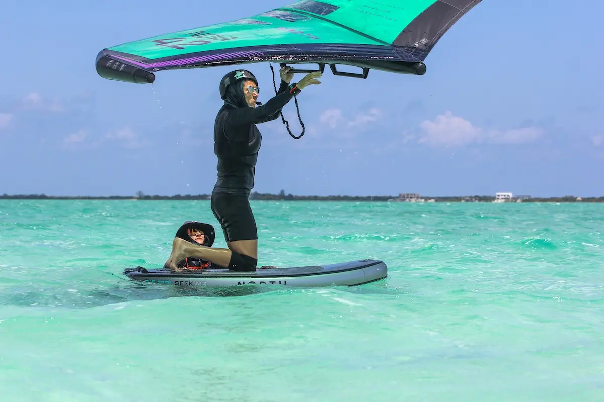 Wingfoil instructor standing on a North Seek Air board holding a teal wing while a student lies on the board in turquoise Belize water during a women's wingfoil retreat.