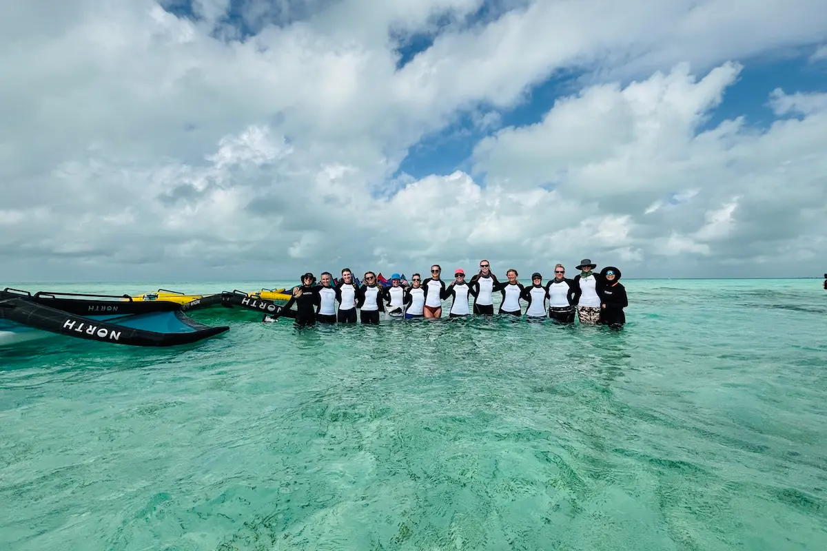 Large group of women in Kite Sisters rash guards standing together in turquoise shallow water with North wings beside them during a women's wingfoil retreat in Belize.