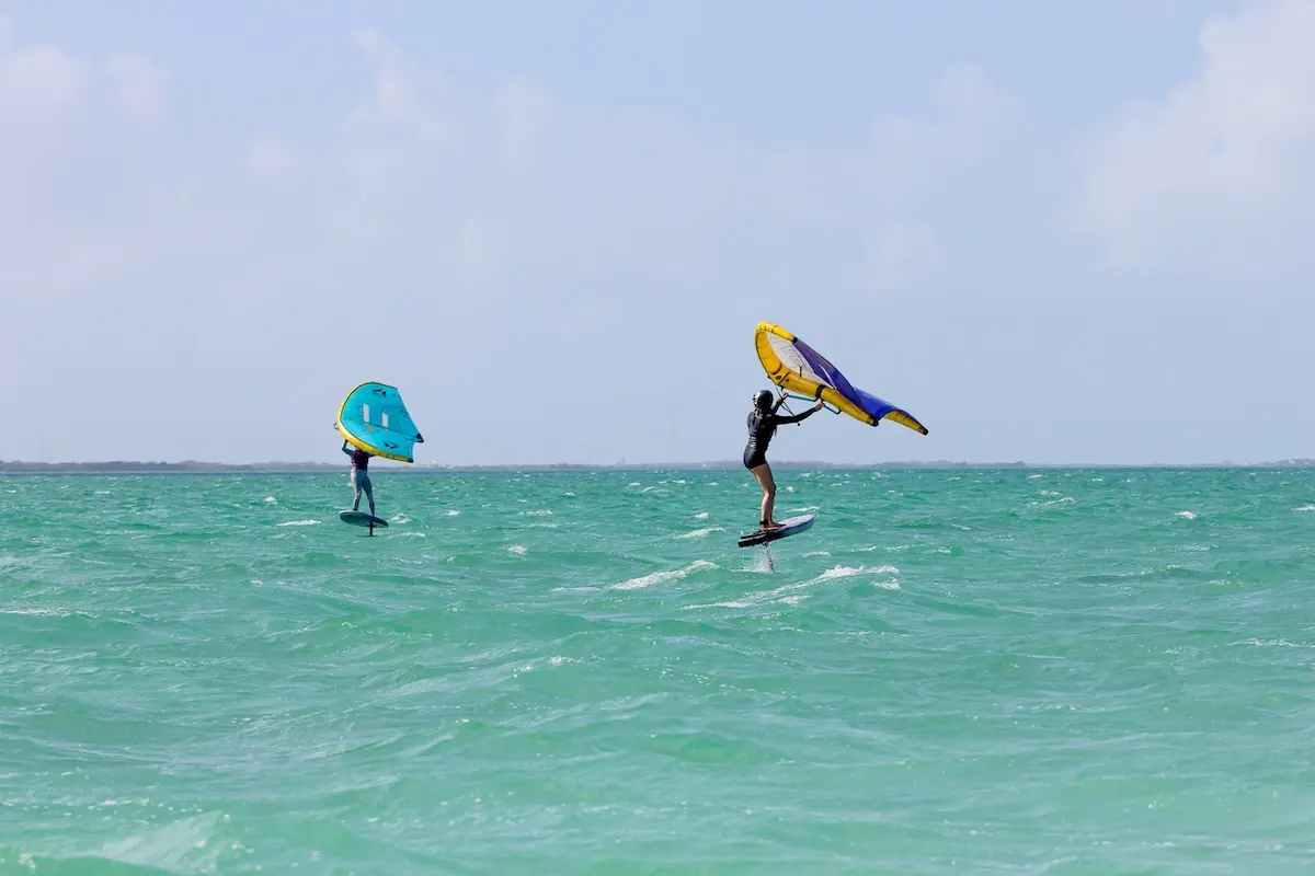 Two women wingfoiling side by side on foil boards with colorful wings over turquoise Belize water during a women's wingfoil retreat in Belize.