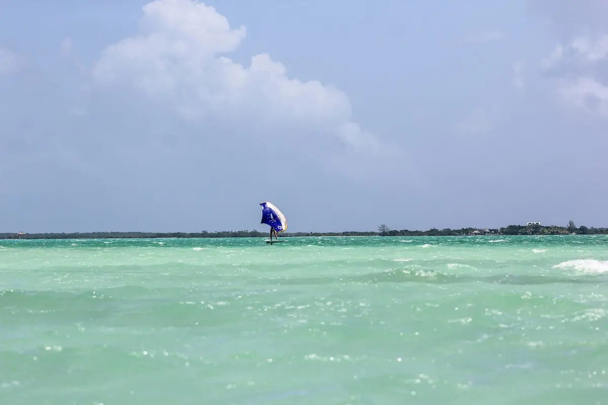 Distant view of a woman wingfoiling on a foil board with a blue wing on the wide turquoise Belize ocean during a women's wingfoil retreat.