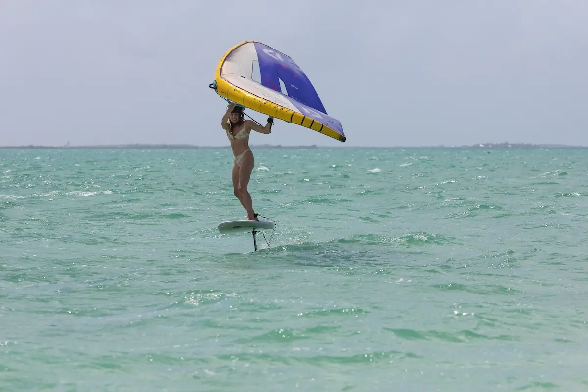 Woman in a bikini foiling above turquoise water holding a yellow and blue wing during a women's wingfoil retreat in Belize.