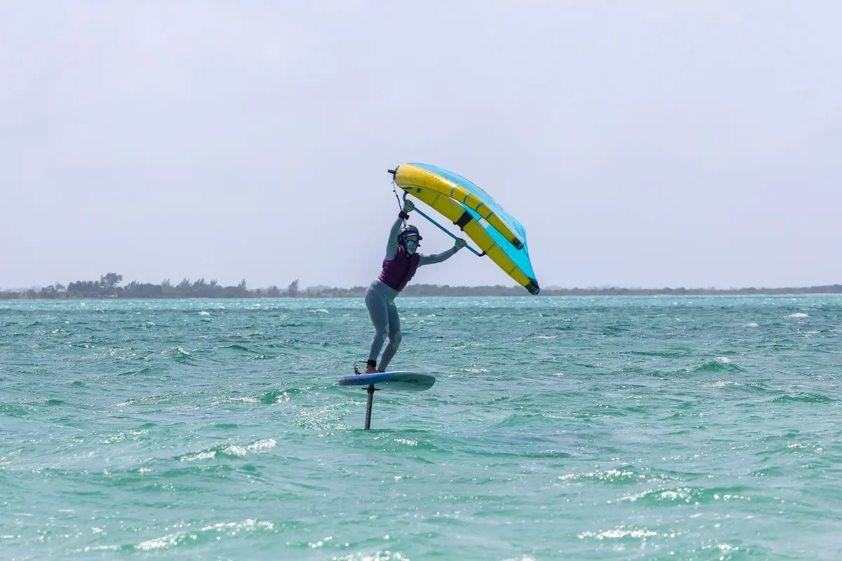 Woman in a wetsuit foiling above the water holding a yellow and blue wing on choppy turquoise Belize water during a women's wingfoil retreat in Belize.