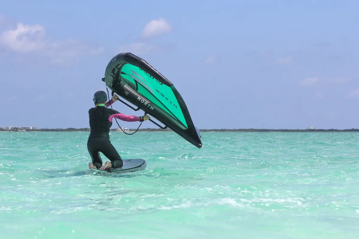 Woman kneeling on a North foil board holding a green North Nova wing on turquoise water during a women's wingfoil retreat in Belize.